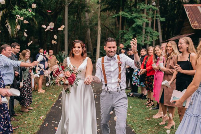 A bride and groom walk down the ailse at their outdoor wedding at The Acre. Guests throw confetti.