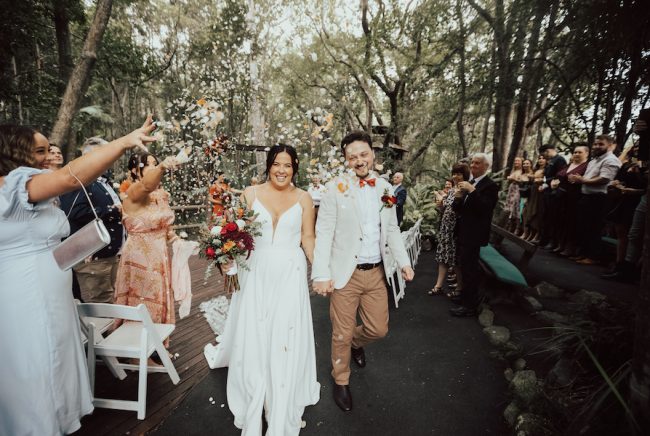 Guests throw confetti over a bride and groom. There are huge trees and nature surrounding them at The Acre Boomerang Farm.